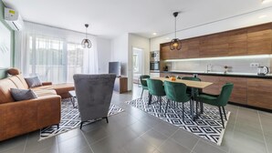 A modern kitchen with wooden cabinets, a dining area with green chairs, and a living room with a brown sofa.