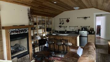 A cozy living room with a fireplace, a wooden ceiling, and a kitchen area with a refrigerator and sink.