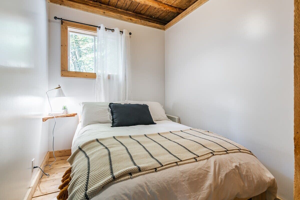Un dormitorio con cama, mesita de noche, lámpara, ventana con cortinas y techo de madera.