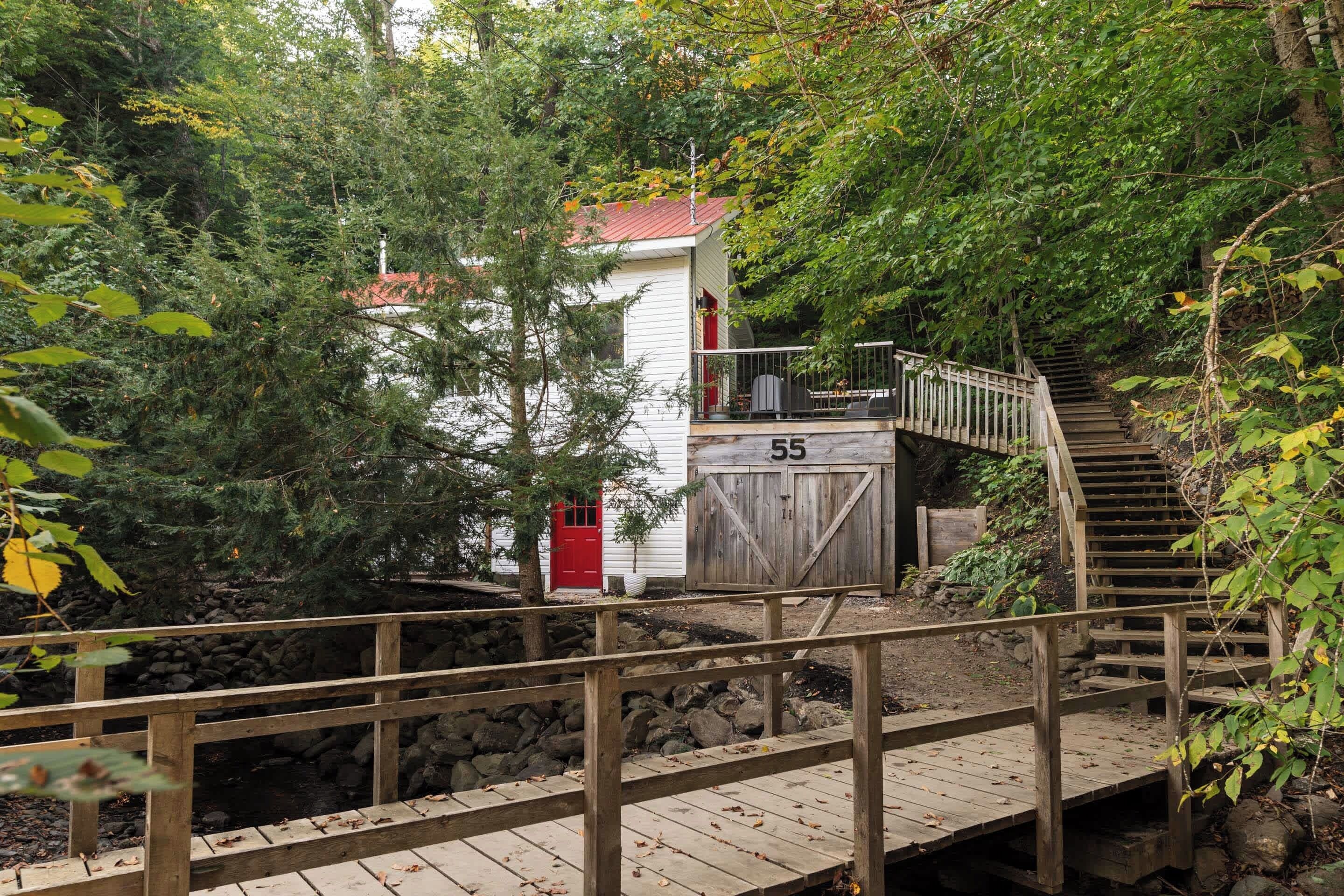 Un pont de fusta que condueix a un edifici blanc amb una porta vermella, envoltat d'arbres frondosos i una paret de pedra.