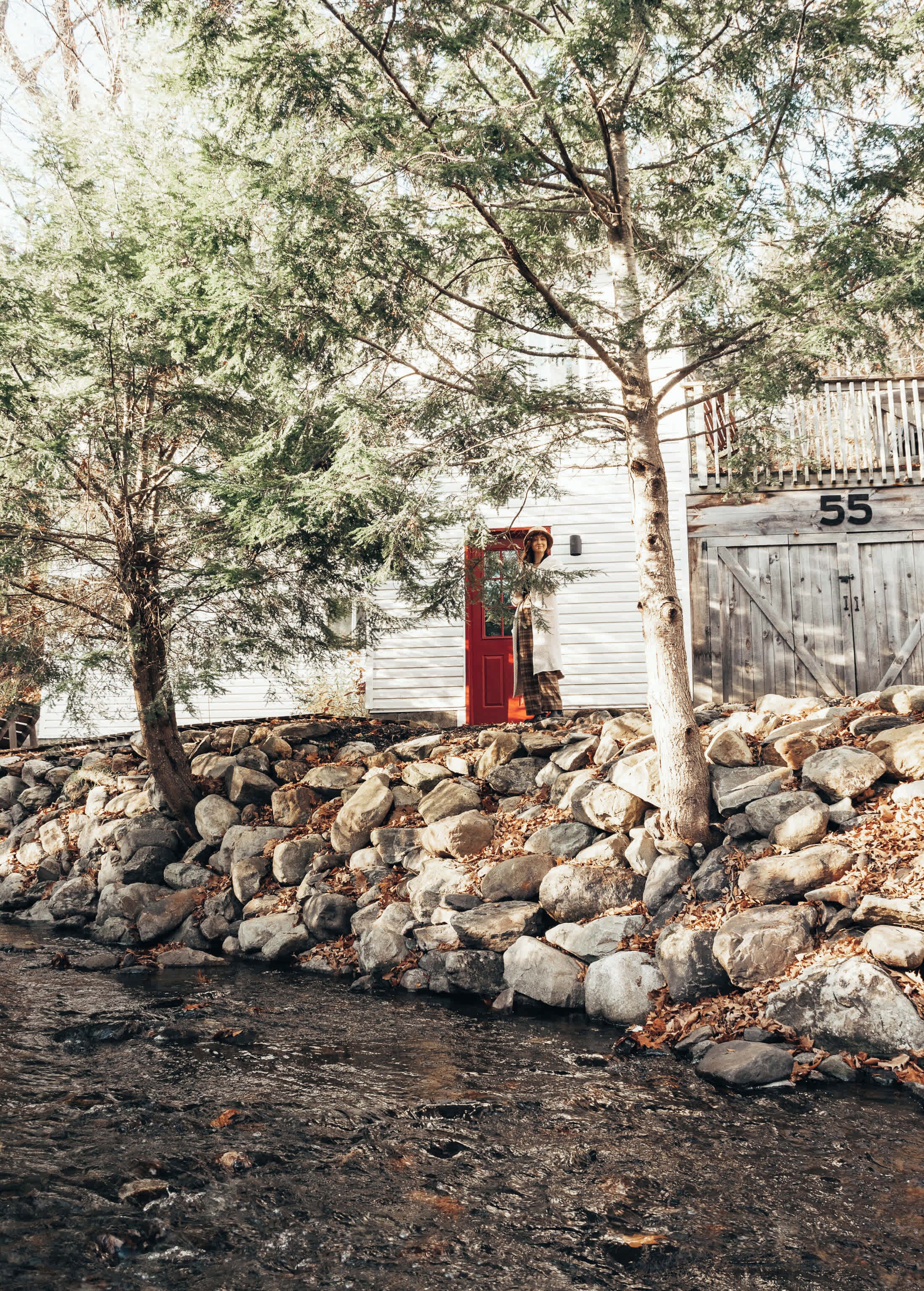 A rustic cabin with a red door, surrounded by a stone wall and trees.