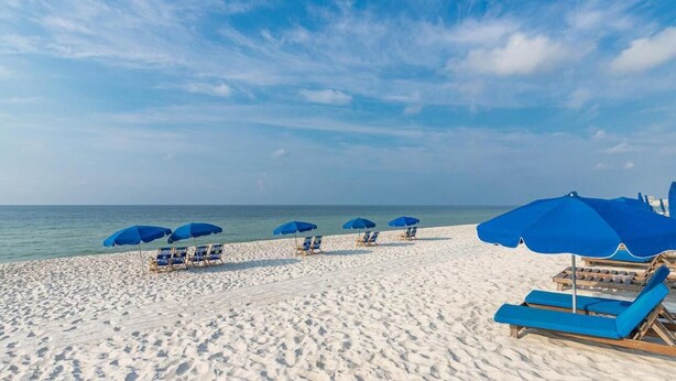 A beach with white sand and a clear blue sky. There are blue umbrellas and lounge chairs along the shore. - Phoenix VII 004 Summer vacation dates available. Just steps to Pool and Beach!! (Orange Beach)