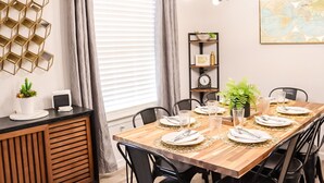 A dining area with a wooden table set for four, black chairs, a black chandelier, a window with blinds, and a decorative wall shelf.