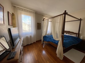 Une chambre à coucher avec un lit à baldaquin, un téléviseur posé sur un meuble en bois et une fenêtre ornée de rideaux légers.