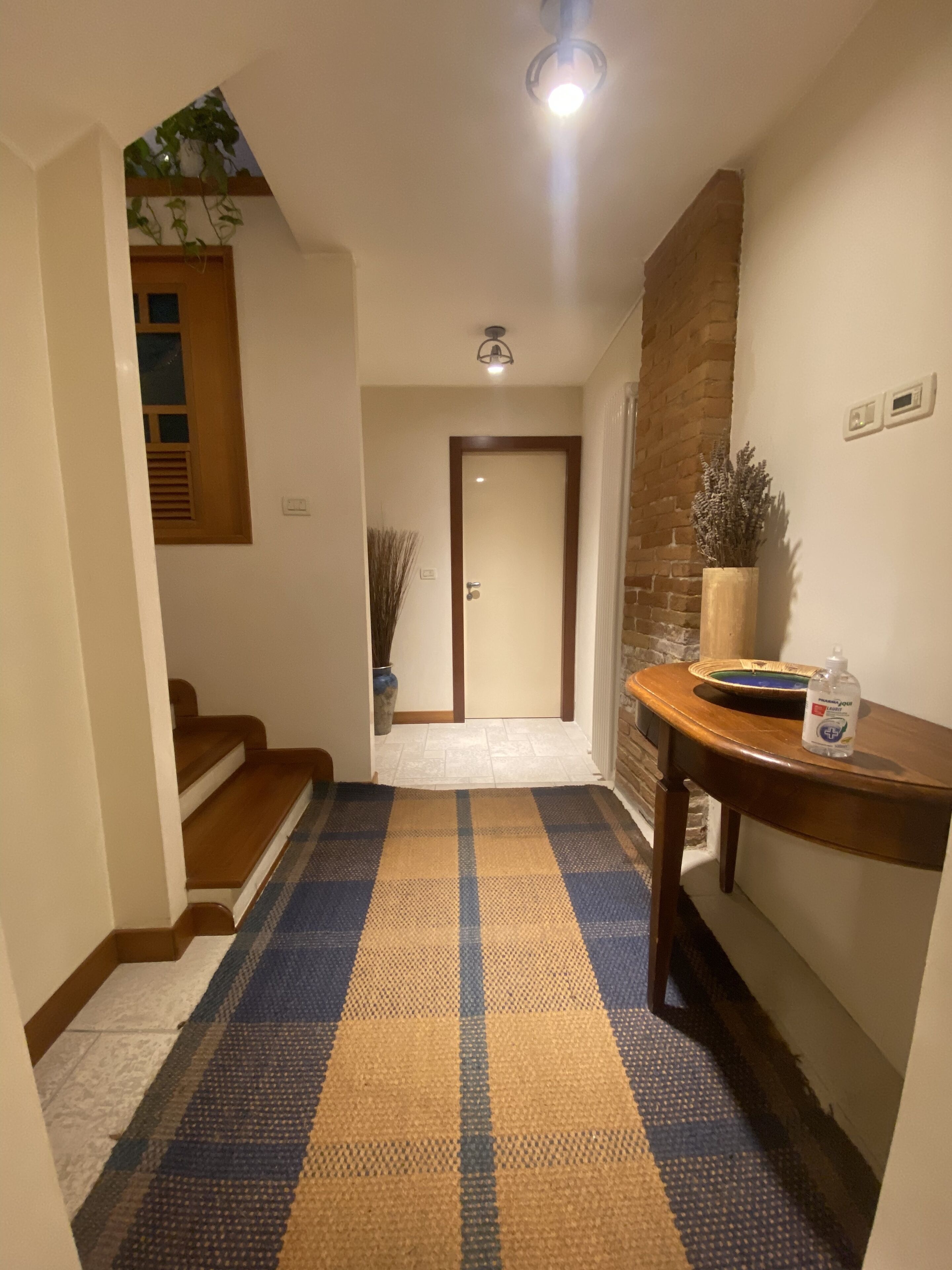 A hallway with a checkered floor, a wooden staircase, a door, and a table with a bottle.