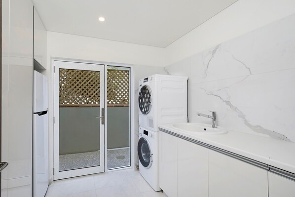 A modern bathroom with a white sink, a built-in washing machine, and a glass door leading to a shower area.