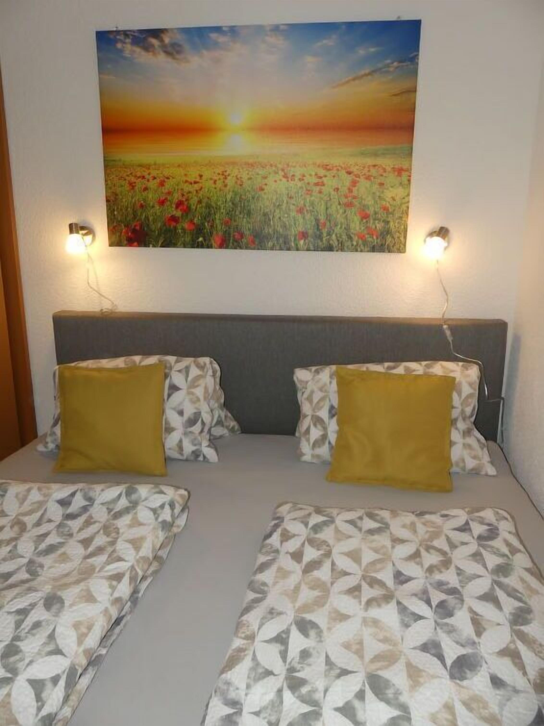 A room with two beds, each with patterned bedding and two yellow pillows. A large painting of a sunset over a field hangs above the beds.