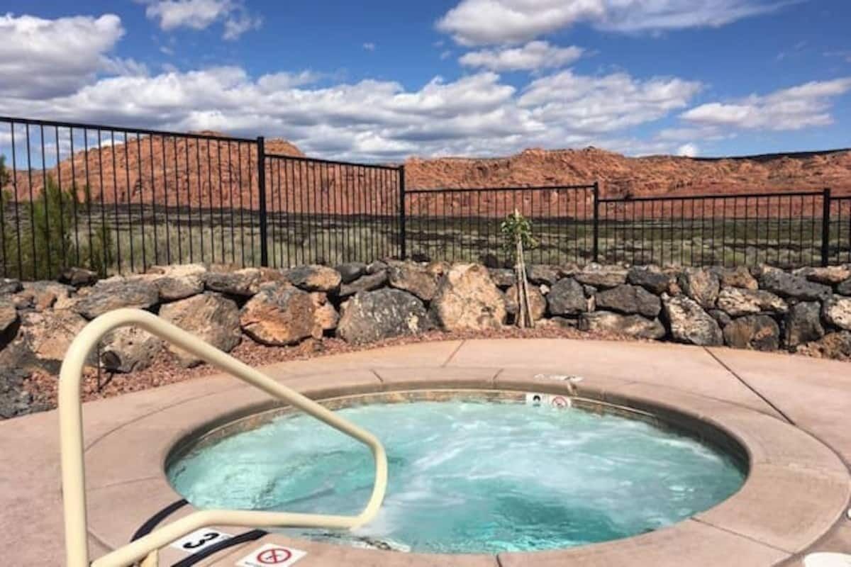 A hot tub with a metal railing, surrounded by a stone wall and a fence, with a mountainous landscape in the background.