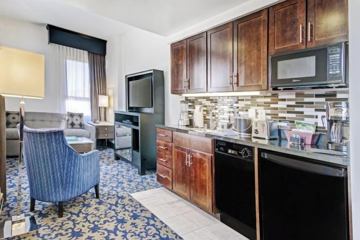 A compact hotel room with a kitchenette, a television, a sofa, and a patterned floor.