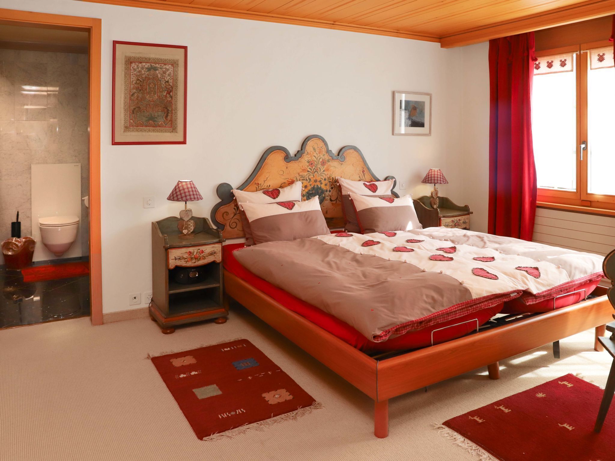 Un dormitorio con una cama de madera, dos mesitas de noche con lámparas, una alfombra roja y blanca estampada, y una ventana con cortinas rojas.