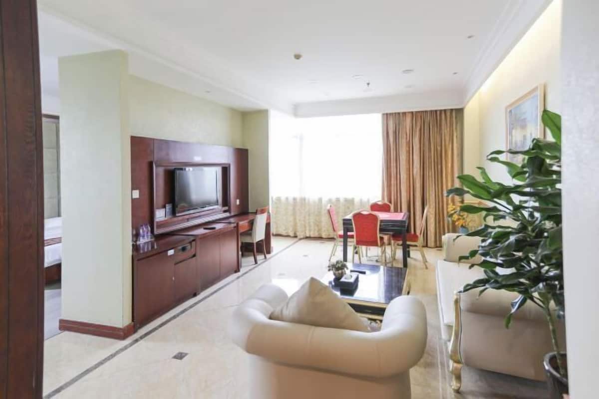 A modern hotel room with a flat-screen TV, a dining area with a red table and chairs, a sofa, and a large potted plant.