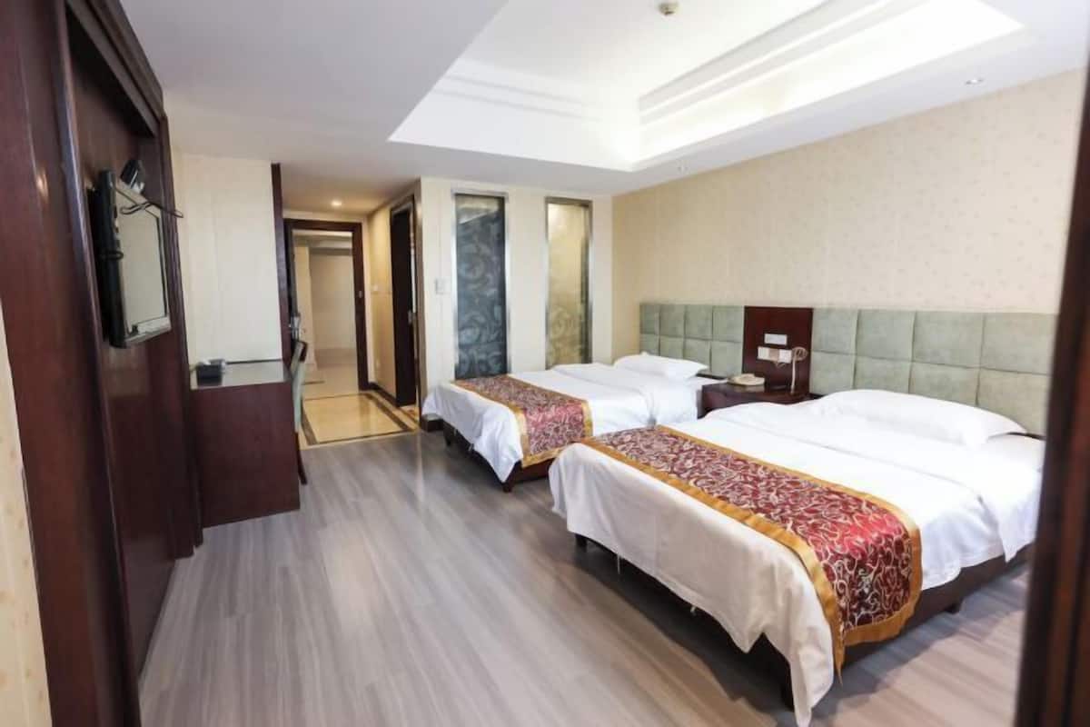 A hotel room with two beds, a wooden wardrobe, a mirror, and a hallway leading to another room.