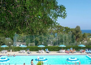 A swimming pool with lounge chairs and umbrellas, surrounded by greenery and a clear sky.