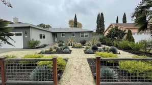 Ein Wohnhaus mit gepflasterter Auffahrt, die zu einer Garage führt, umgeben von begrünten Gärten mit verschiedenen Pflanzen und einem Metallzaun.