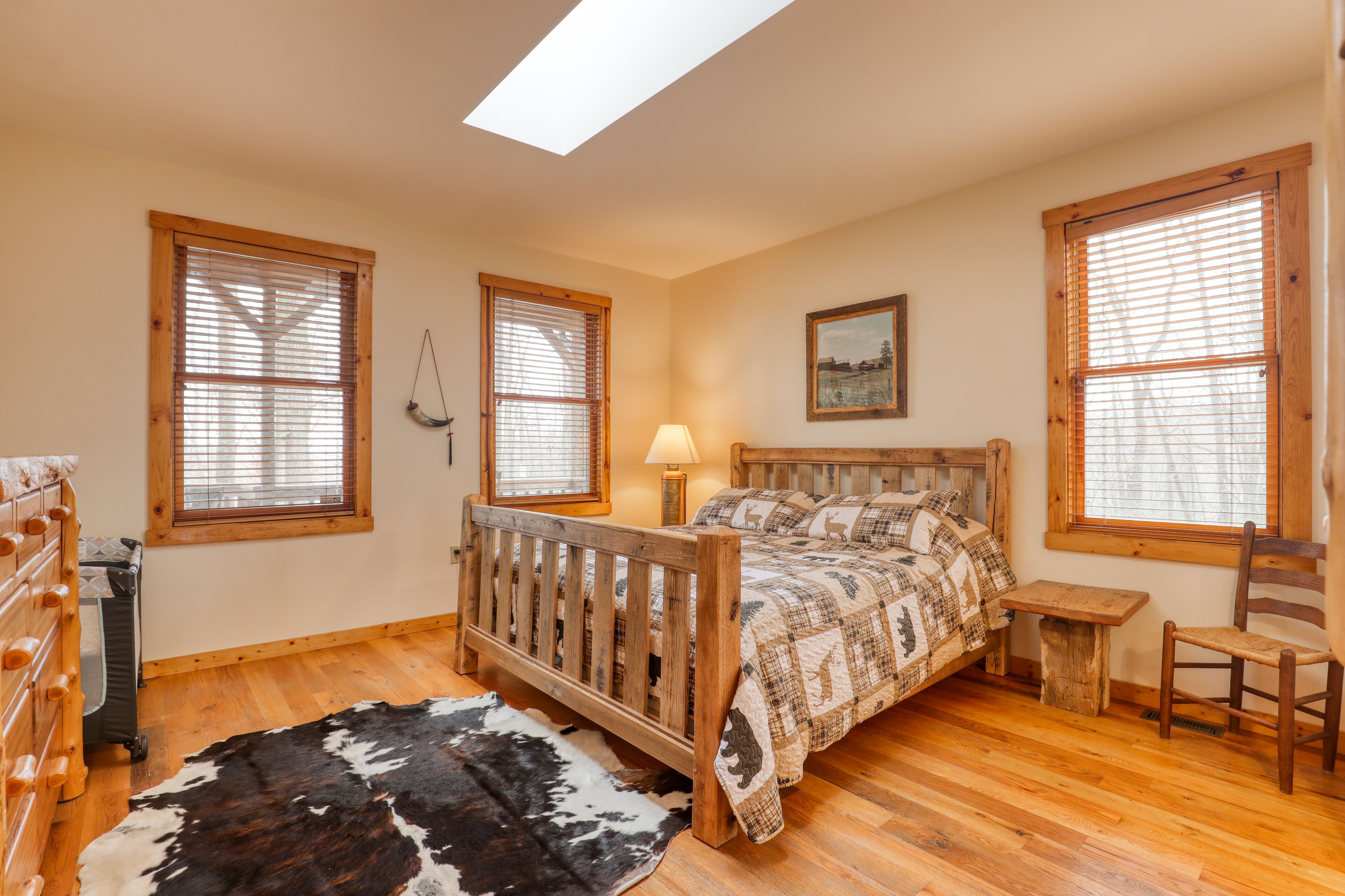 Quarto com cama de madeira, mesa de cabeceira, cadeira, tapete de couro e amplas janelas com persianas.