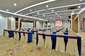 Uma sala de conferências com fileiras de mesas e cadeiras, um palco e uma tela de projeção.