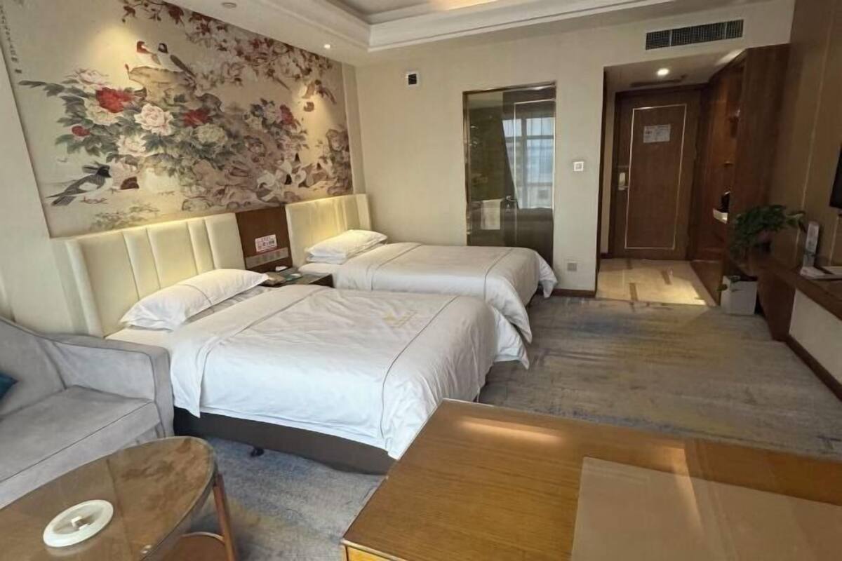 Habitación de hotel con dos camas, un sofá, una mesa de centro de madera, una pared decorativa con motivos florales y una puerta que da al baño.