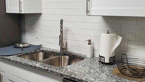 A kitchen with two stainless steel sinks, a granite countertop, a paper towel dispenser, and a black metal rack. - Coastal Home w/ Outdoor Living, 10 min to Beach (Vero Beach)