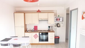 A modern kitchen with white cabinets, a red hanging light, a microwave, and a coffee maker. There is a dining table with chairs and a trash bin. - DINARD - LA RICHARDAIS T2- NEAR SEA AND RANCE (La Richardais)