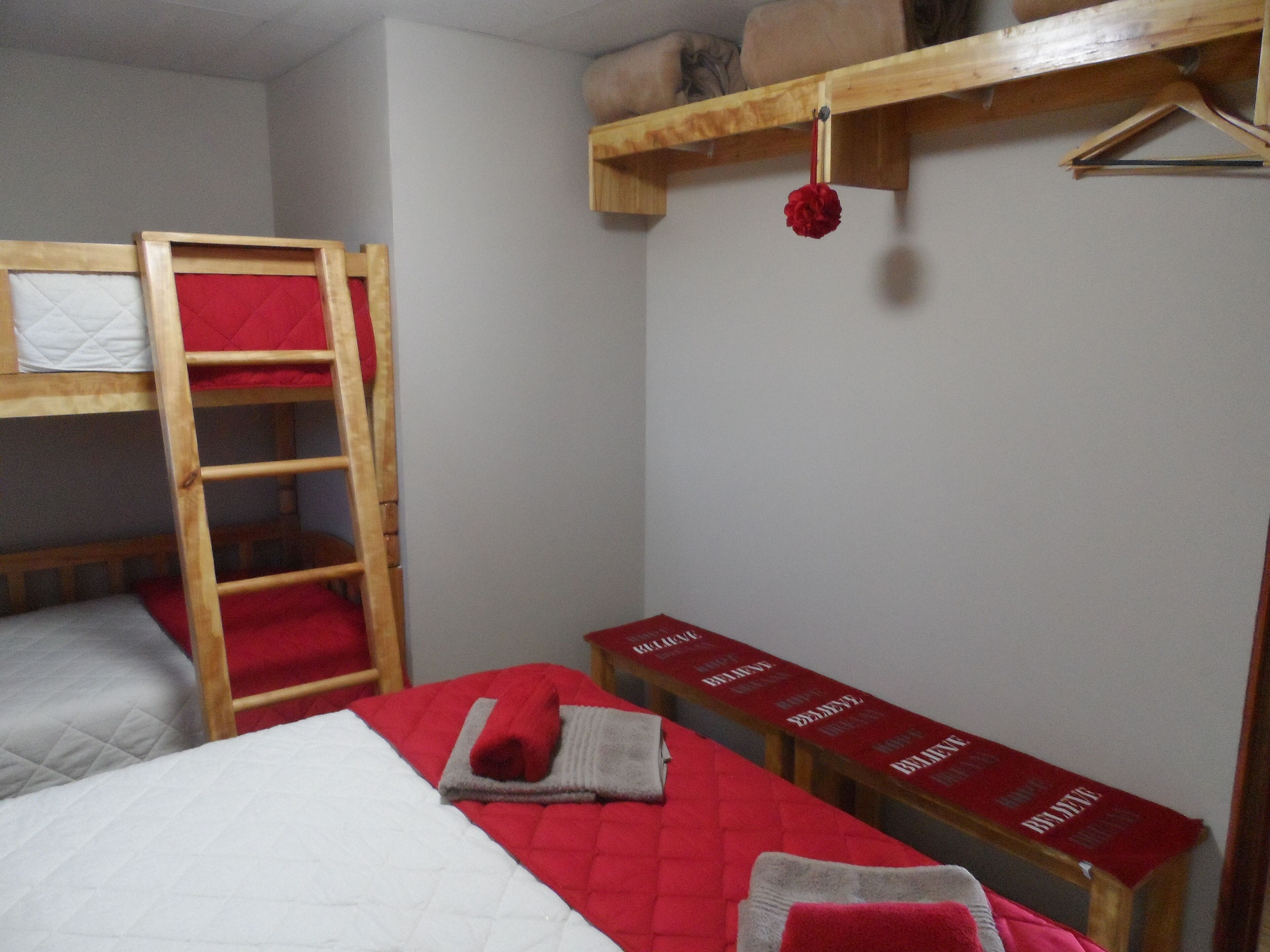A dormitory room with bunk beds, a wooden ladder, a red quilted bedspread, and a shelf with a red cushion and a hanging ornament.