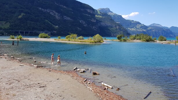 A beach with clear water, a bridge in the background, and mountains. - Two-bedroom Apartment for 4-5 Guests With Lake and Mountain Views (Emmetten)