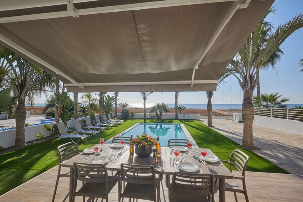 A covered outdoor dining area with a pool, chairs, and a table set for a meal.