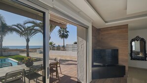A modern living room with a TV, wooden panel wall, and a glass door leading to a pool area with outdoor furniture. - Ayia Napa Holiday Villa Sv9 (Sotira)