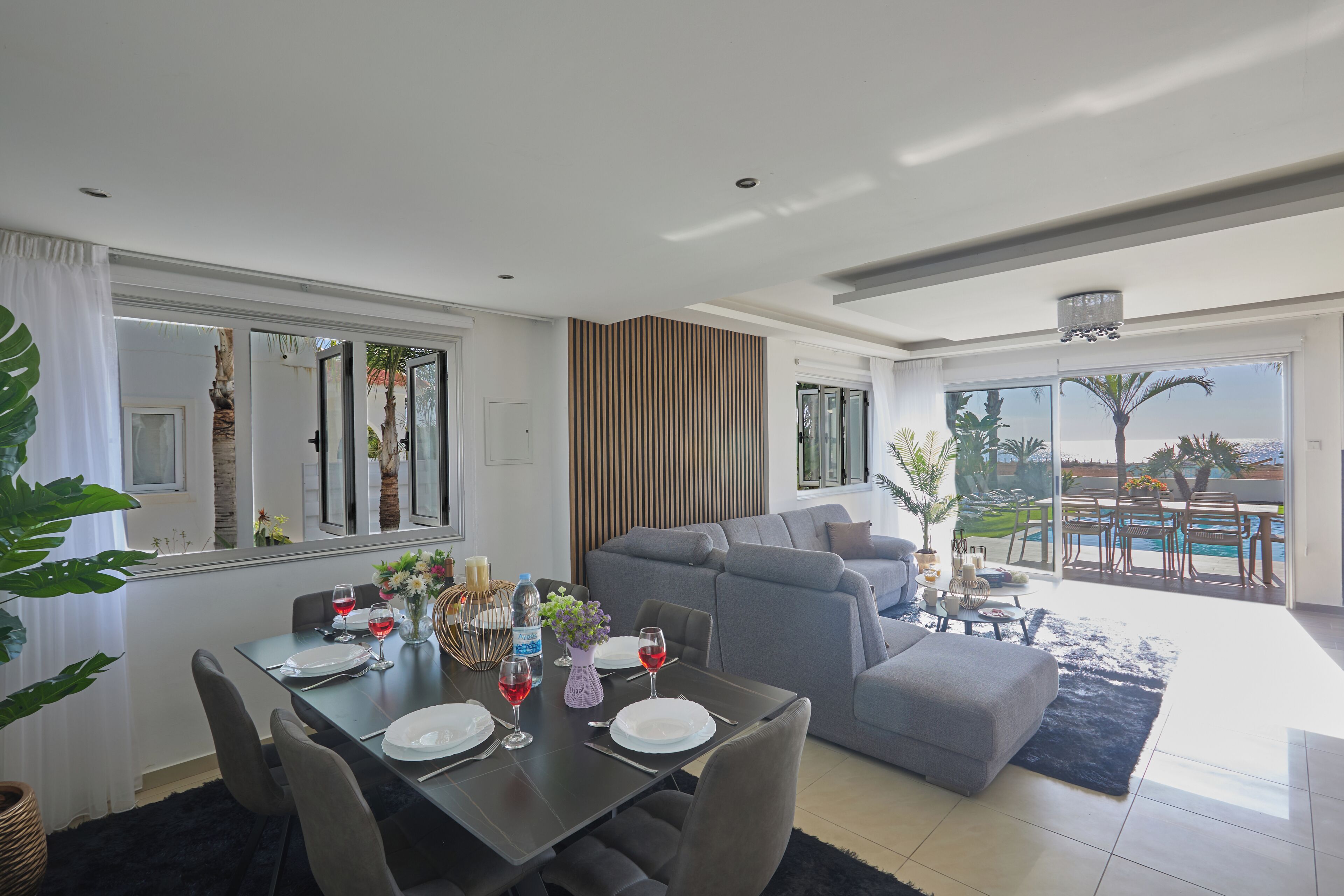A modern living room with a dining area, grey sofas, and a view of the pool.