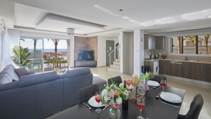 A modern living room with a sofa, dining table set for two, and a kitchen area visible through an open door. - Ayia Napa Holiday Villa Sv9 (Sotira)