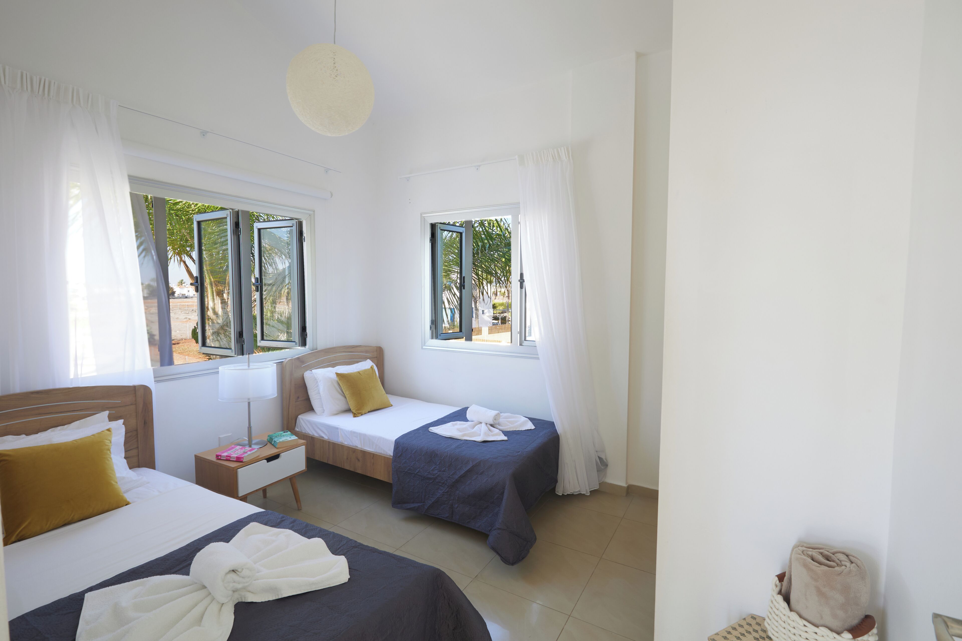 A hotel room with two beds, a nightstand, a lamp, and a view of palm trees.