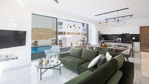 Una sala de estar moderna con un sofá verde, una mesa de centro y un televisor. Se observa un comedor con una mesa y sillas, y una cocina con encimera negra. Se ve una piscina a través de una puerta de vidrio.