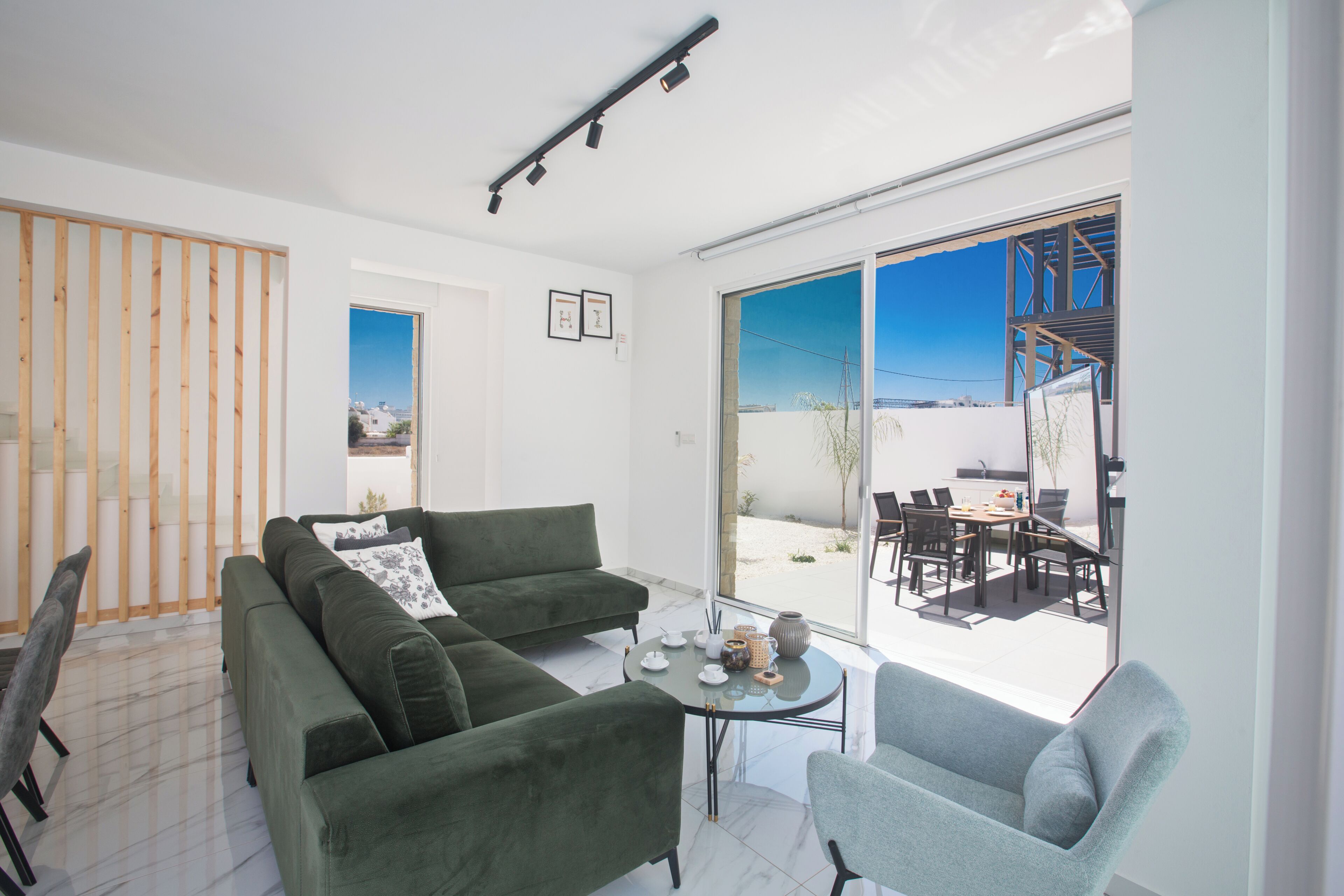 A modern living room with a green sofa, a grey armchair, and a small table with tea set. Large windows offer a view of the outside.
