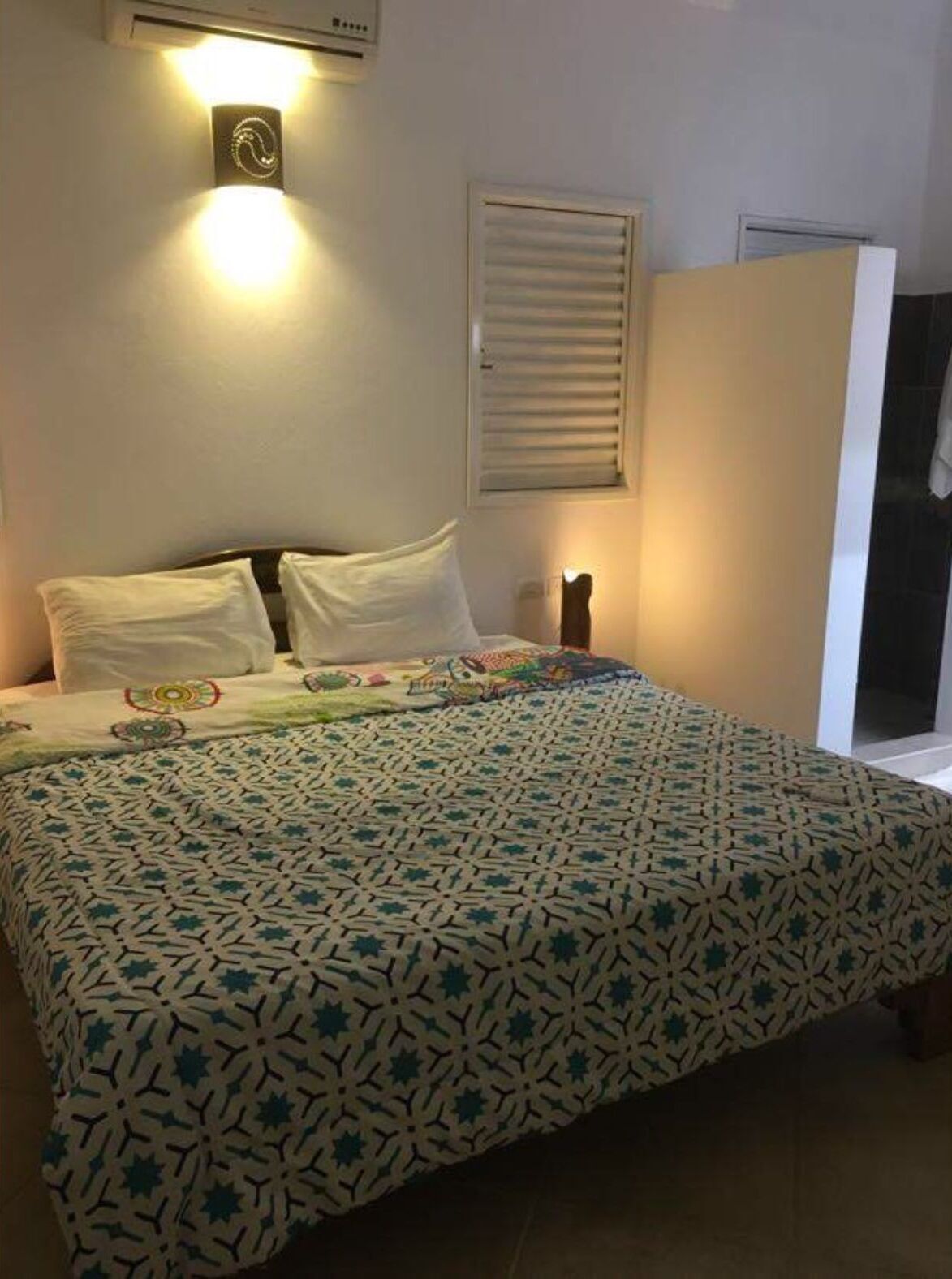 Una cama bien hecha con una colcha estampada, una lámpara de pared y una ventana con persianas.