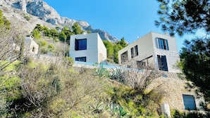 Un edificio moderno de dos pisos con fachada de piedra, ubicado en una ladera con cielo azul despejado.
