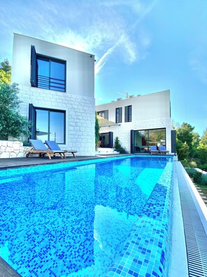 A modern poolside area with a clear blue pool, lounge chairs, and a two-story building with large windows and a stone facade.