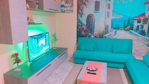 A living room with a teal sofa, a flat-screen TV on a stand, a coffee table with a flower, and a wall mural.