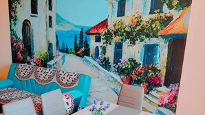 A dining area with a table set for four, surrounded by chairs. A painting of a coastal scene with buildings and flowers is on the wall behind the table.