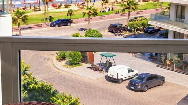 A view from a balcony overlooking a beach with palm trees, a parking area, and a building with a glass facade. - Modern Chalet With Garden Views and Shared Pool in Port Said (Al-Manasrah)