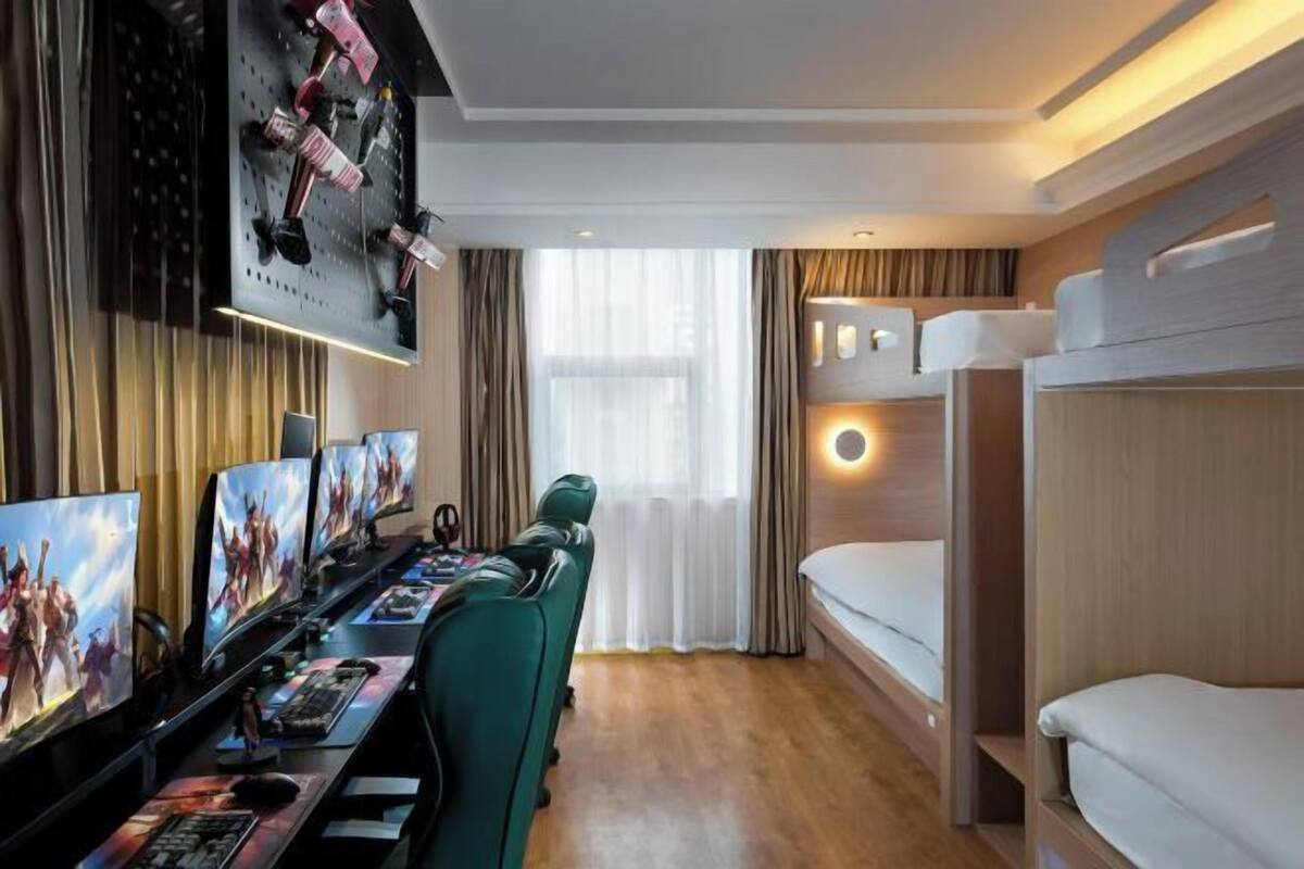 Une chambre d’hôtel équipée d’un bureau, de plusieurs écrans d’ordinateur, d’une chaise verte, d’un lit superposé et d’une grande fenêtre avec des rideaux.