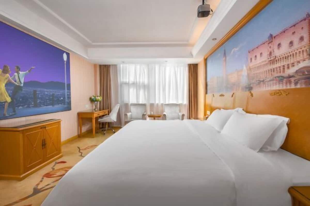 Une chambre d’hôtel avec un grand lit, une télévision, un bureau avec une chaise et une fenêtre avec des rideaux.