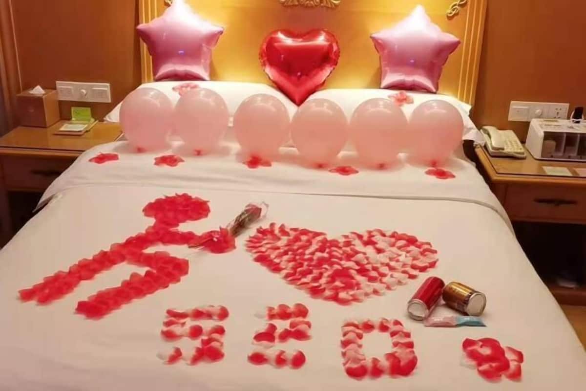 Une chambre d’hôtel avec un lit décoré de pétales de rose formant les mots « Je t’aime », des ballons roses et un ballon en forme de cœur.