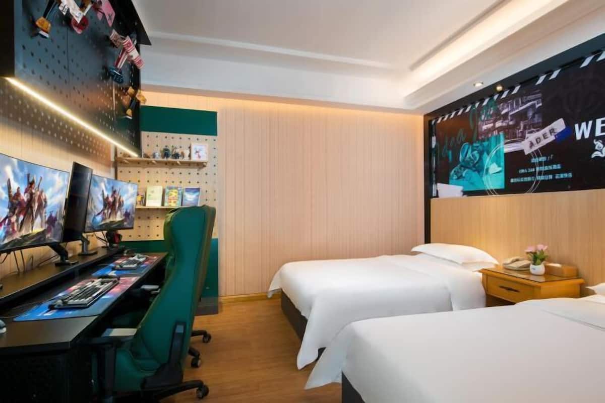 Une chambre d’hôtel équipée d’un espace de jeu vidéo, avec deux lits, un bureau doté d’écrans et une grande affiche accrochée au mur.