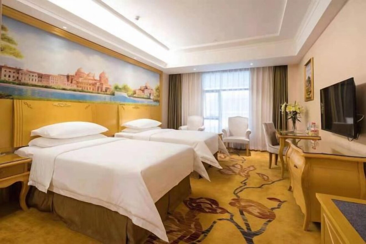 A hotel room with two beds, a desk, a chair, a TV, and a painting on the wall.