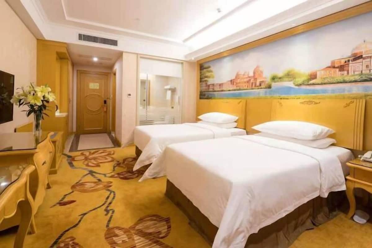Une chambre d’hôtel avec deux lits, un bureau, une chaise, un tableau au mur et un tapis à motifs floraux.