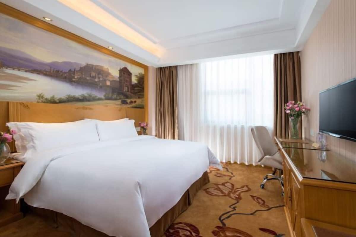 Une chambre d’hôtel avec un grand lit, un bureau avec une télévision, une fenêtre avec des rideaux et un tableau au mur.