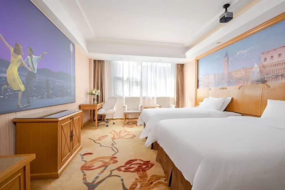 Une chambre d’hôtel avec deux lits, un bureau, une chaise, une télévision et une grande fenêtre avec des rideaux.