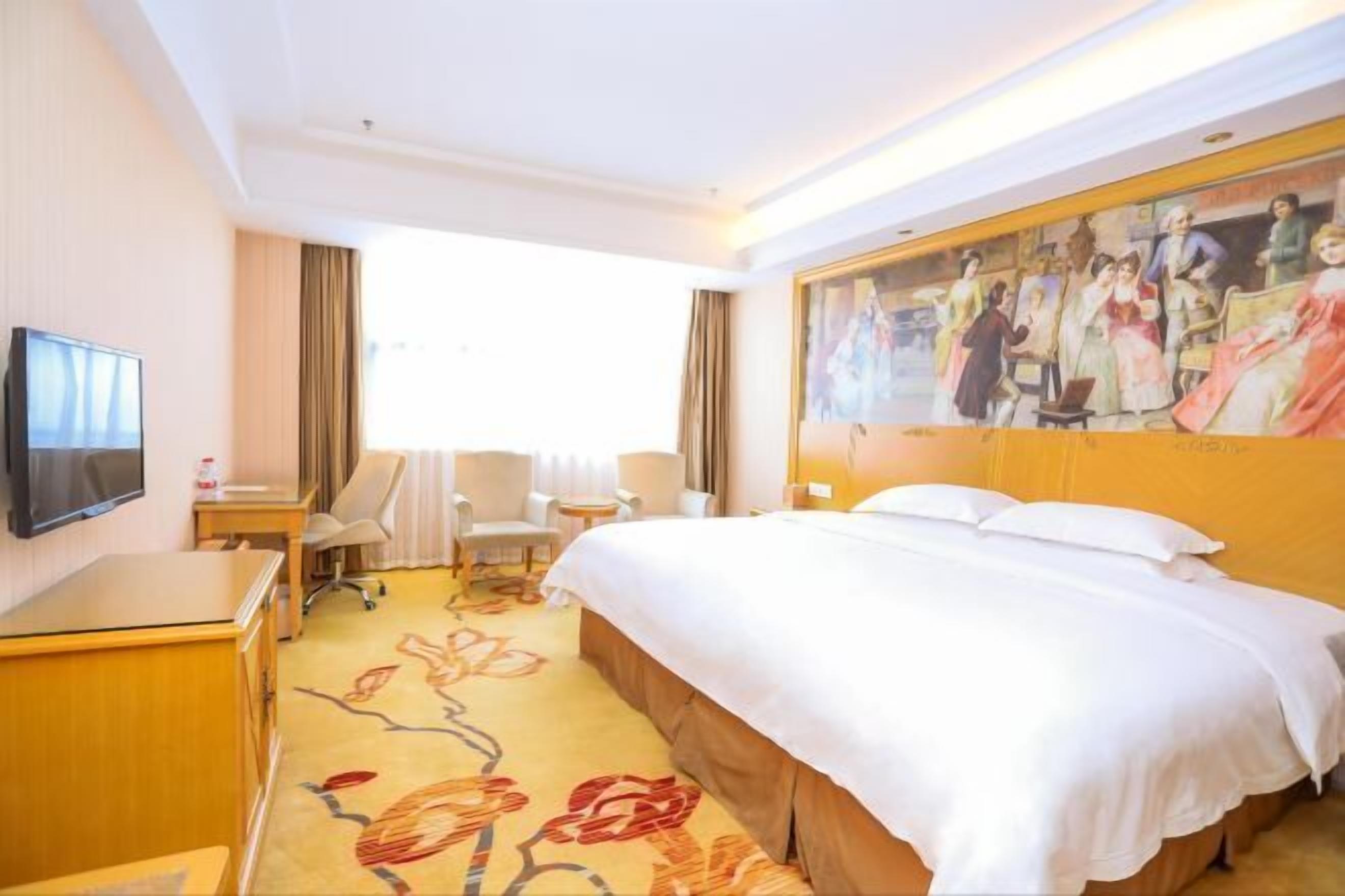 A hotel room with a bed, a desk, a chair, a TV, and a painting on the wall.