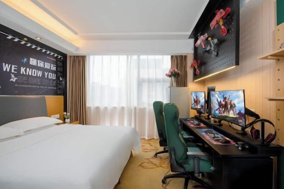 Une chambre d’hôtel avec un lit, un bureau doté de deux écrans, des chaises vertes et un mur orné d’une affiche motivante.