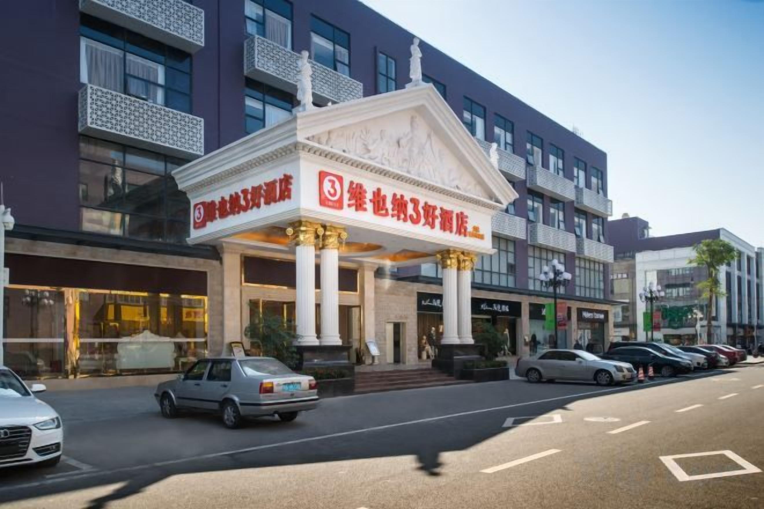 Un hotel con una entrada destacada, un estacionamiento con varios autos y una calle con edificios al fondo.