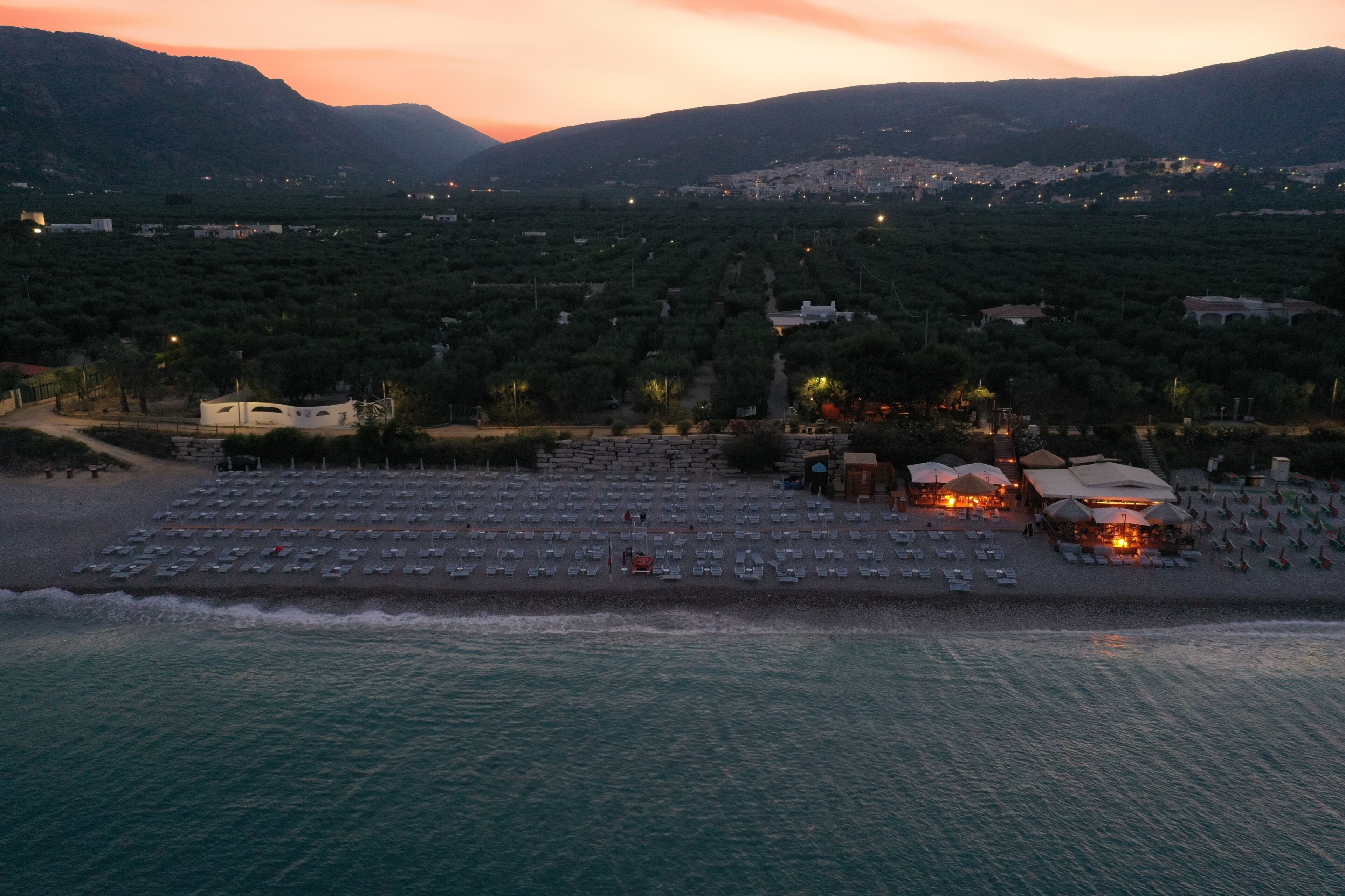 A coastal area with a beach, a row of beach umbrellas, and a building with a roof.